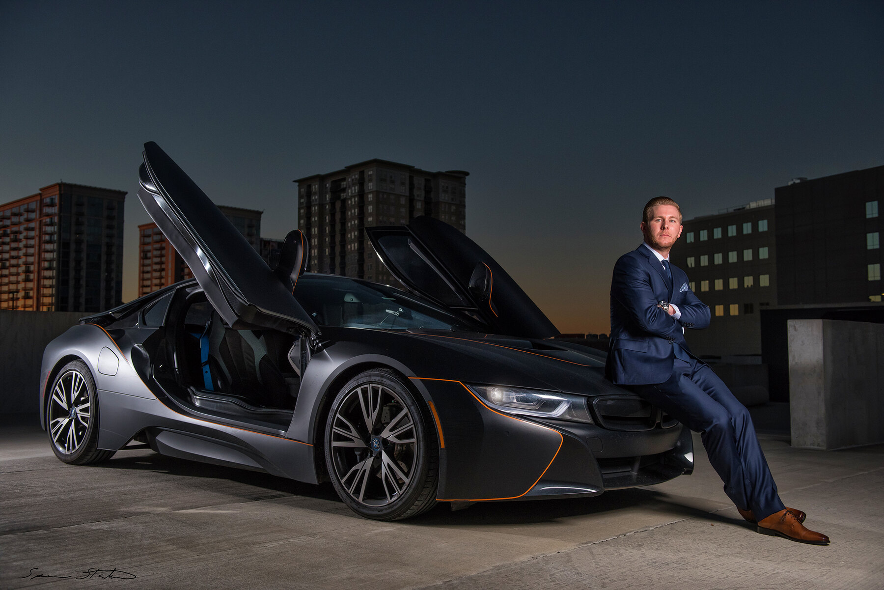 Entrepreneur Ryan White, founder of Social Revelation, seated beside a BMW i8 with its doors raised on a city rooftop at dusk