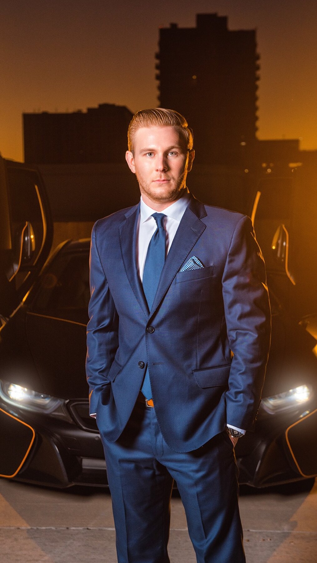 Entrepreneur Ryan White, founder of Social Revelation, in a blue suit with hands in pockets, a BMW i8 with headlights on behind him