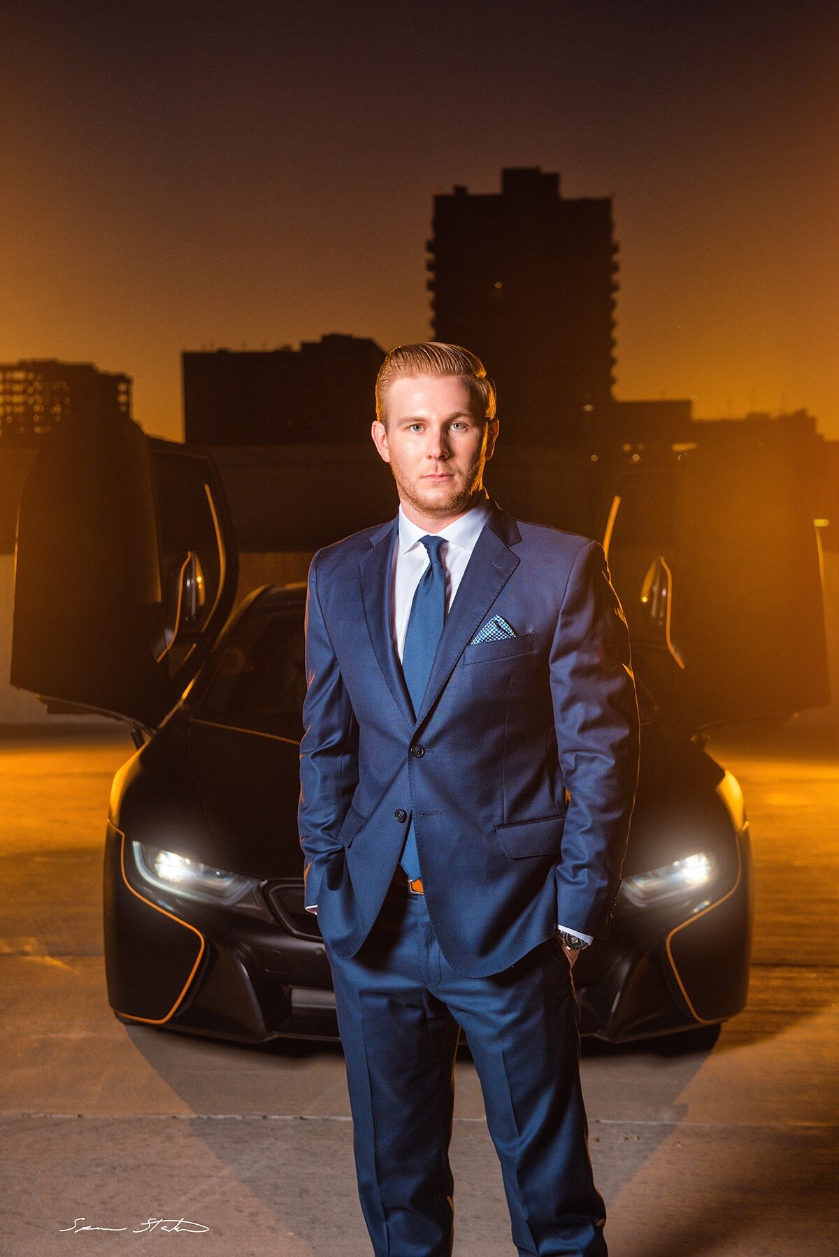Entrepreneur Ryan White, founder of Social Revelation, in a blue suit standing in front of a BMW i8 on a rooftop at sunset
