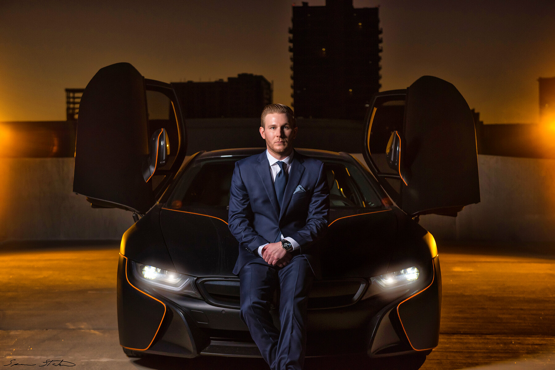 Entrepreneur Ryan White, founder of Social Revelation, seated on the front of a BMW i8 with its doors raised at golden hour