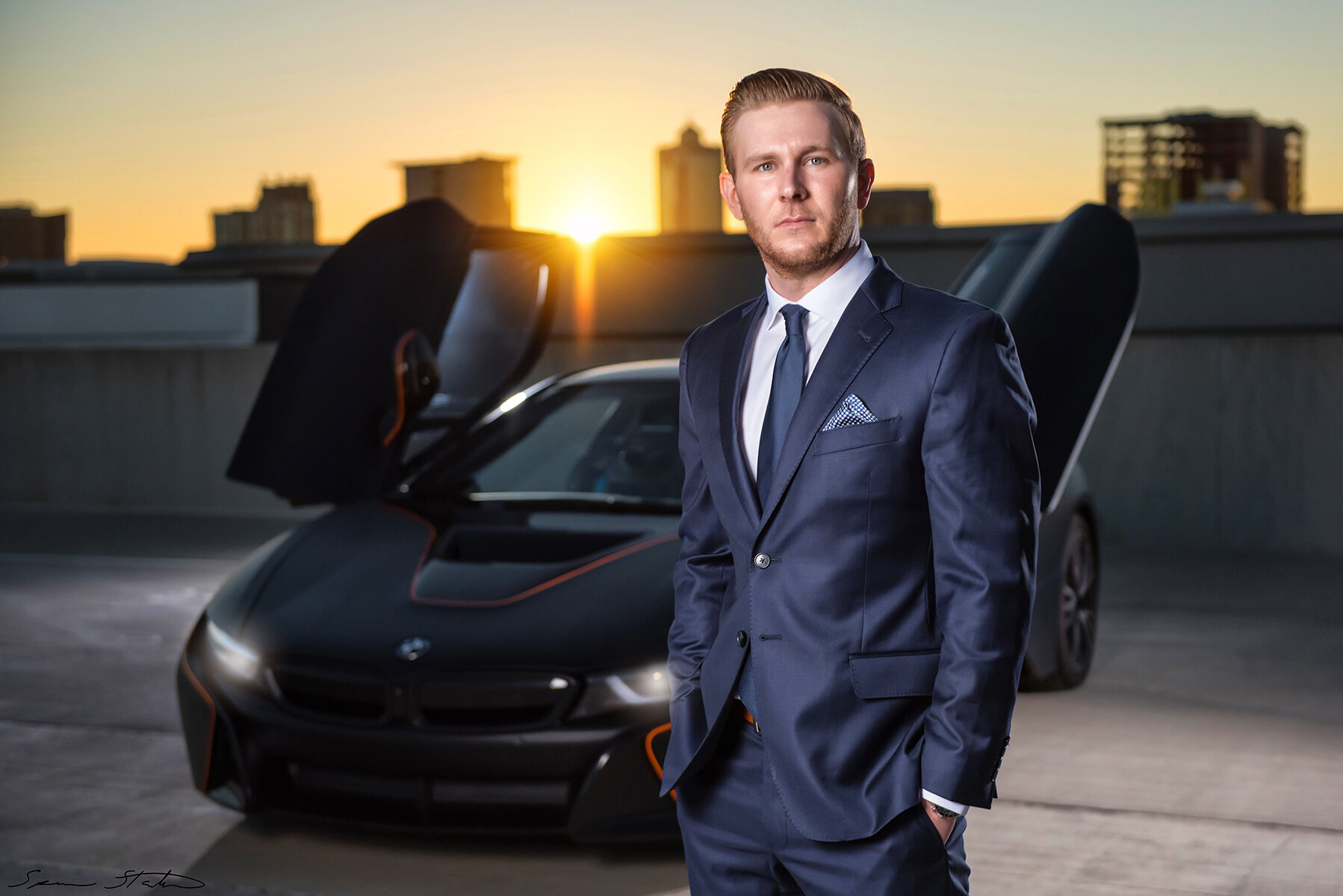 Entrepreneur Ryan White, founder of Social Revelation, in a navy suit beside a BMW i8 with the sun setting over the skyline