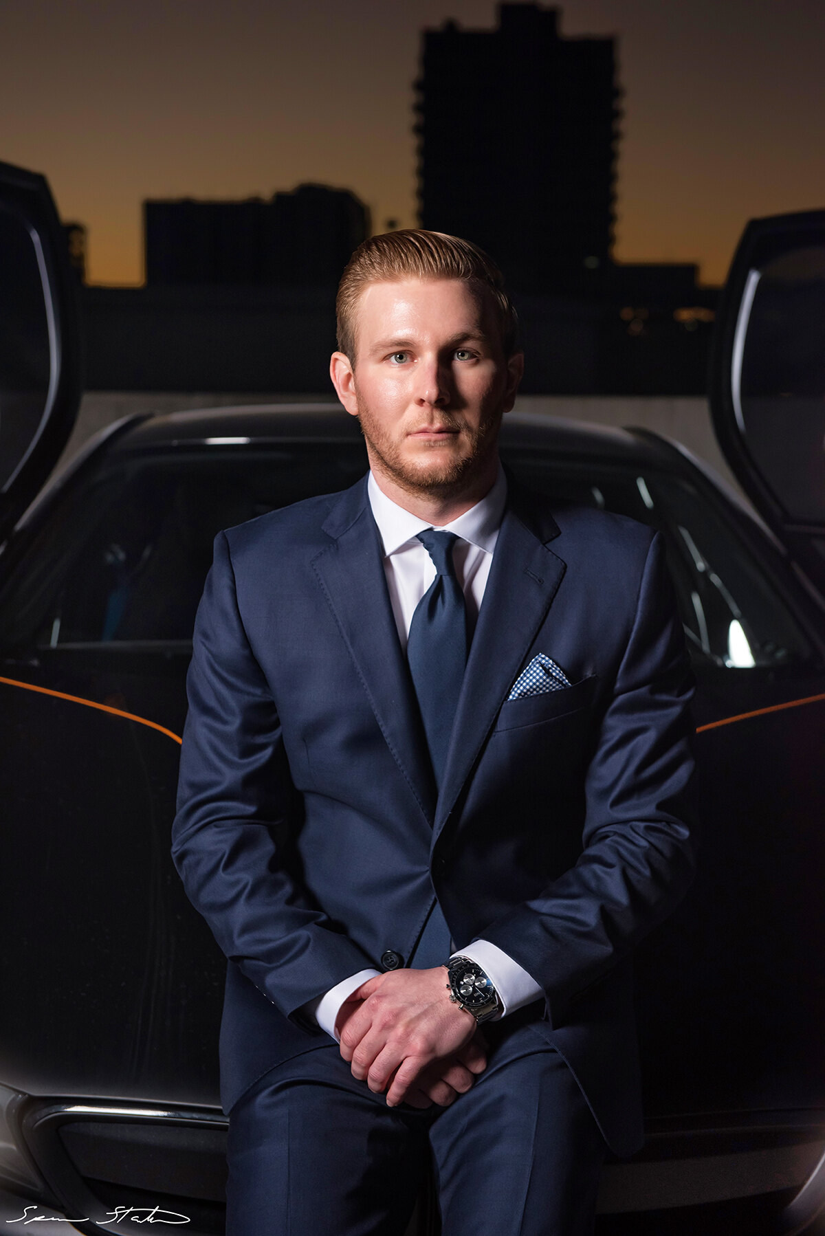 Entrepreneur Ryan White, founder of Social Revelation, seated in a navy suit in front of a BMW i8 with its doors raised at dusk